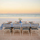 Dining table set for four with a blue tablecloth on a sandy beach at sunset.