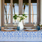 Elegant table setting with a floral tablecloth, plates, glasses, and a vase of flowers in a room with large windows.