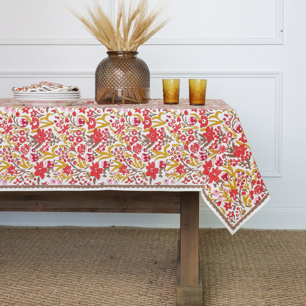 A tablecloth with a red, orange, and pink floral pattern set on a table with two glasses and a vase with wheat on top.