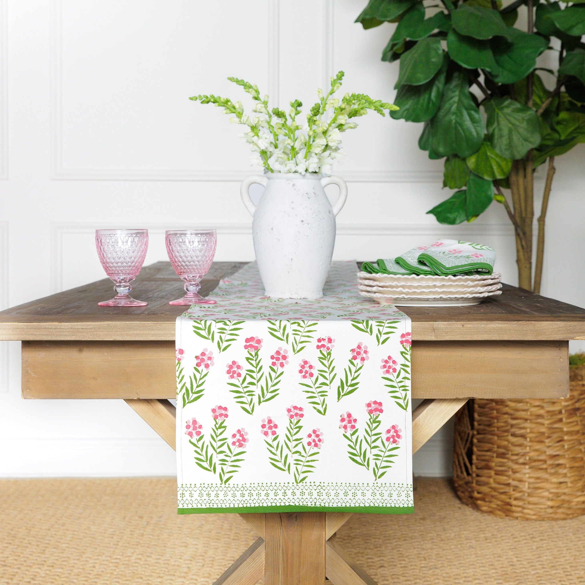 A rectangular table runner with a pink phlox floral pattern displayed on a wooden table. The table is set with pink glasses, a white vase, and greenery in the background.