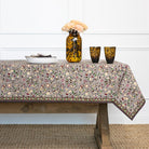 Tablecloth with floral pattern on a wooden table, with a vase and glasses in the background.