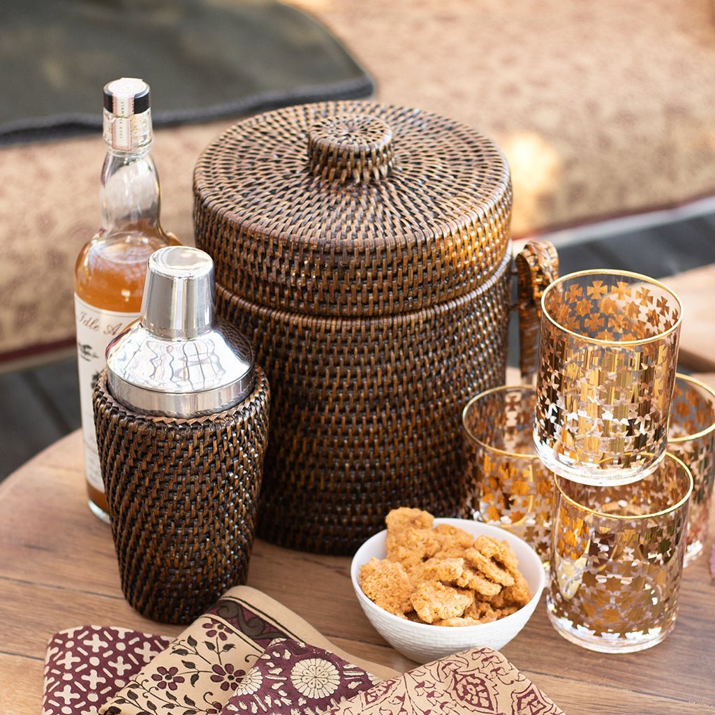 Rattan barware and gold bourbon glasses on a wood table.