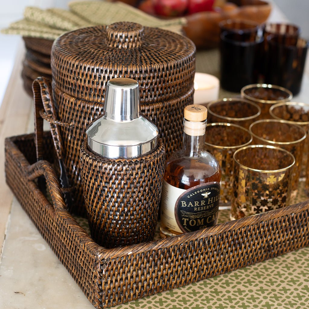 Cocktail shaker, bottle, and glasses on a wicker tray with a candle in the background.