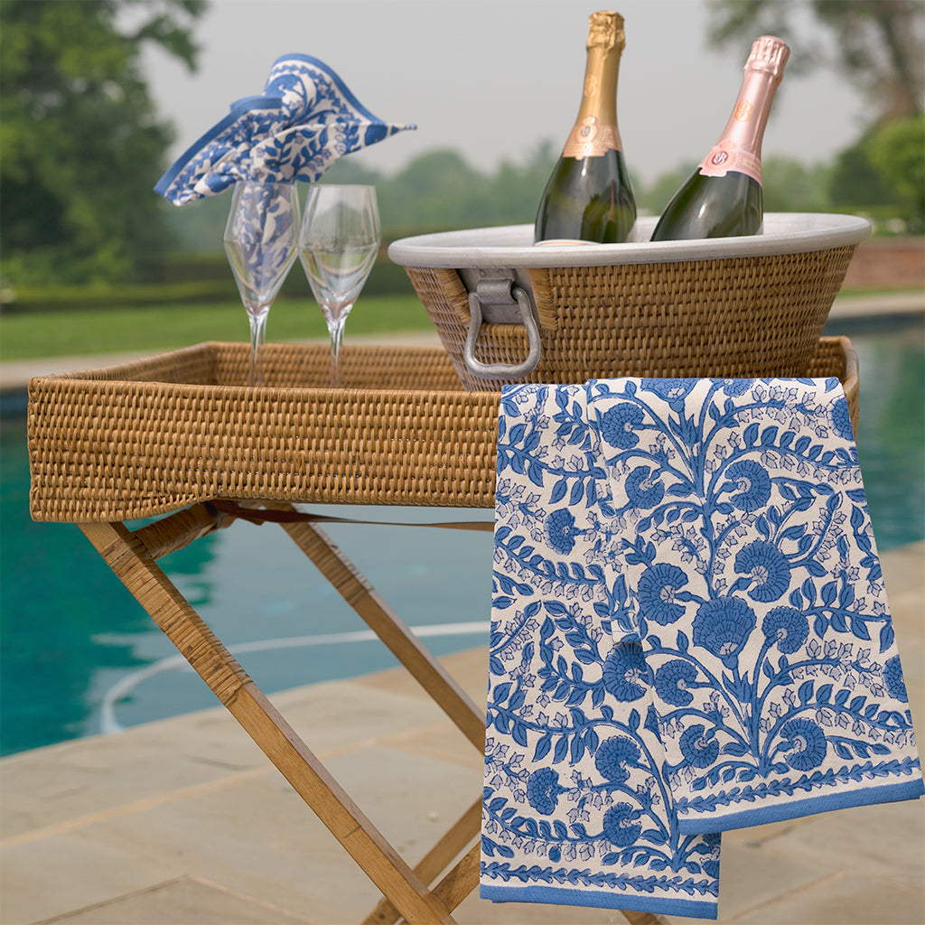 Wicker bar cart with blue patterned towel, champagne bottles, and glasses by a poolside.