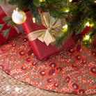 Red gift box with gold bow under a Christmas tree on a decorative tablecloth.