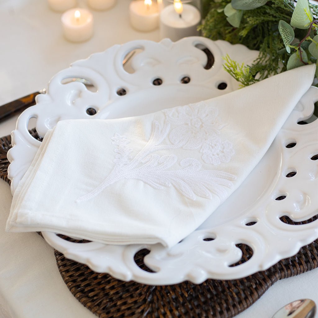 White napkin on a decorative white plate with candles and greenery in the background