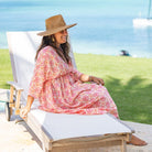 Woman in a floral dress and straw hat sitting on a lounge chair by the water.