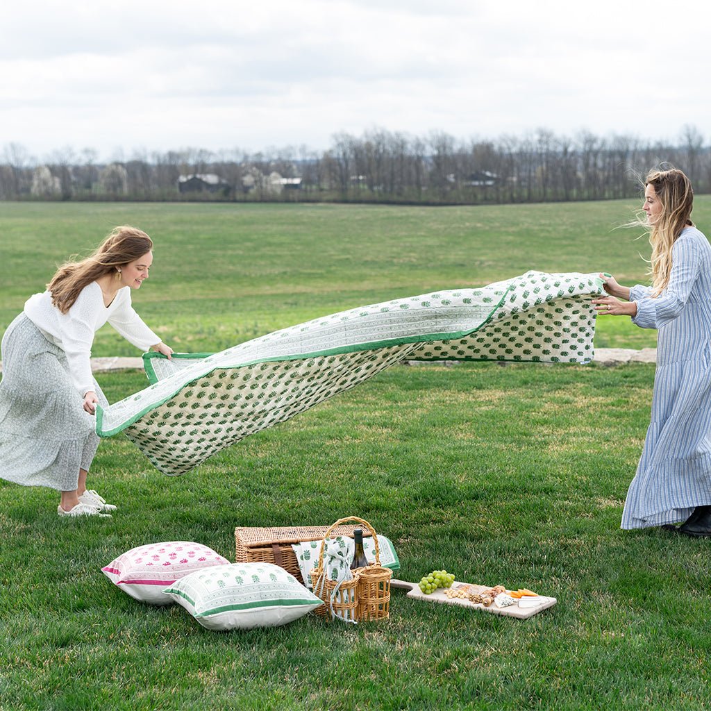 Two women spreading out a picnic blanket on a grassy field with a picnic setup.