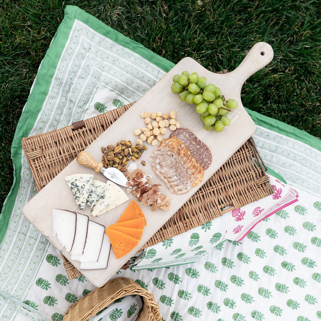 Picnic setup with a cheeseboard, bread, and grapes on a green blanket outdoors.