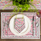 Table setting with a pink decorative placemat, white plate, and silverware on a wooden table.