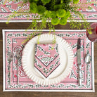 Table setting with a pink decorative placemat, white plate, and silverware on a wooden table.