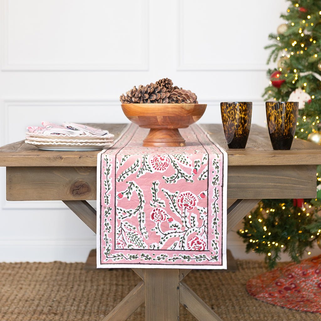 Table with a pink floral table runner, wooden bowl with pinecones, and two dark glass cups on a wooden table.