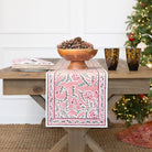 Table with a pink floral table runner, wooden bowl with pinecones, and two dark glass cups on a wooden table.