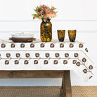 Table setting with a patterned tablecloth, vase with flowers, and glasses on a wooden table.