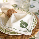 White napkin with green leaf design on a woven plate and tablecloth.