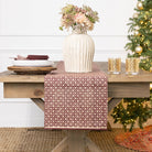 Decorative table setting with a patterned table runner, vase with flowers, and Christmas tree in the background.