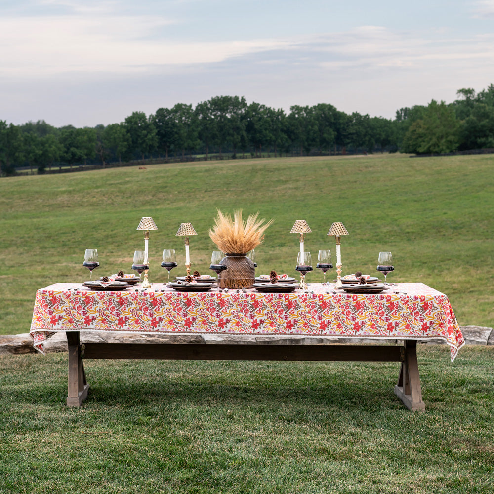 Dining table set with floral tablecloth, plates, glasses, and centerpieces outdoors on grass.