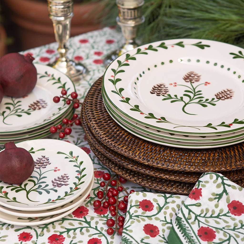 Set of decorative plates with holly and pinecone patterns on a table with red flowers.