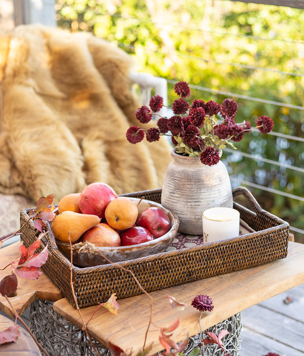 Decorative tray with fruits, a vase of flowers, and a candle on a wooden surface outdoors.