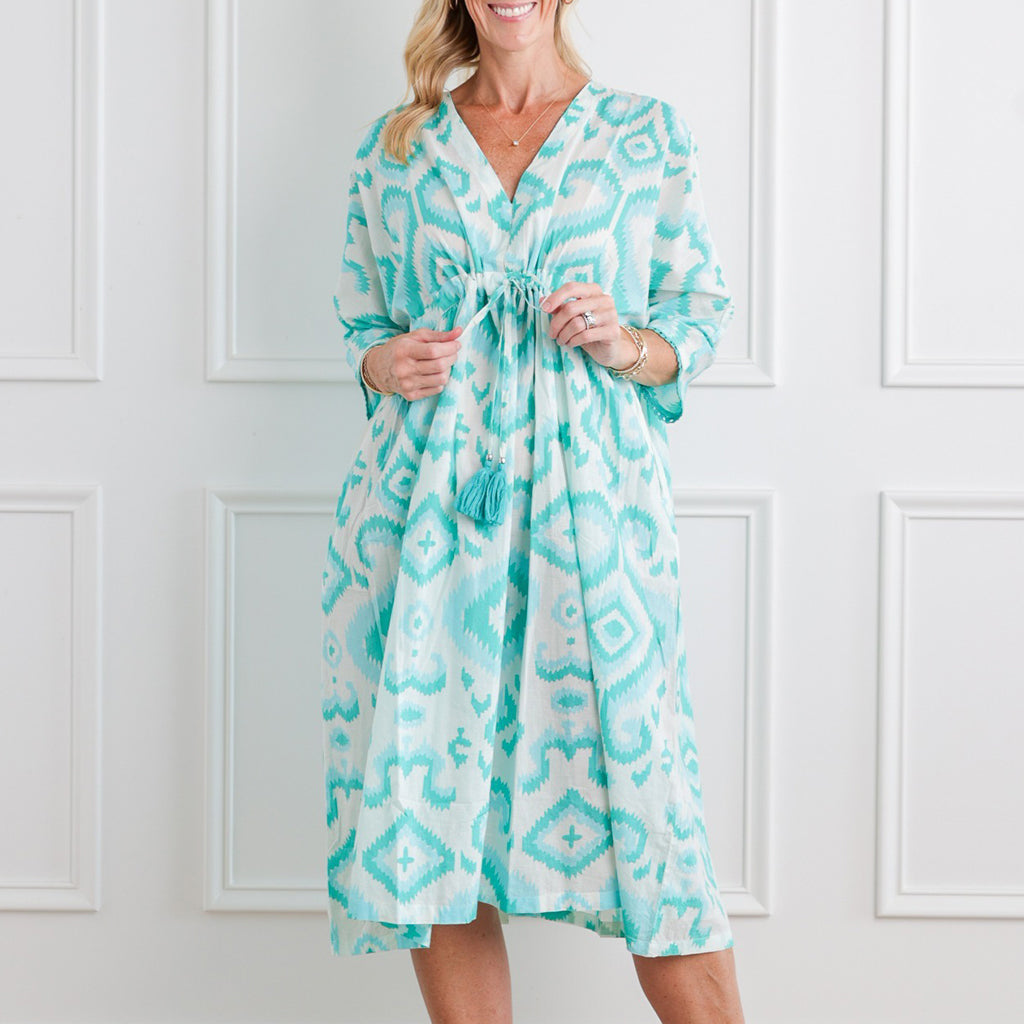 Woman wearing a turquoise patterned dress against a white paneled wall.