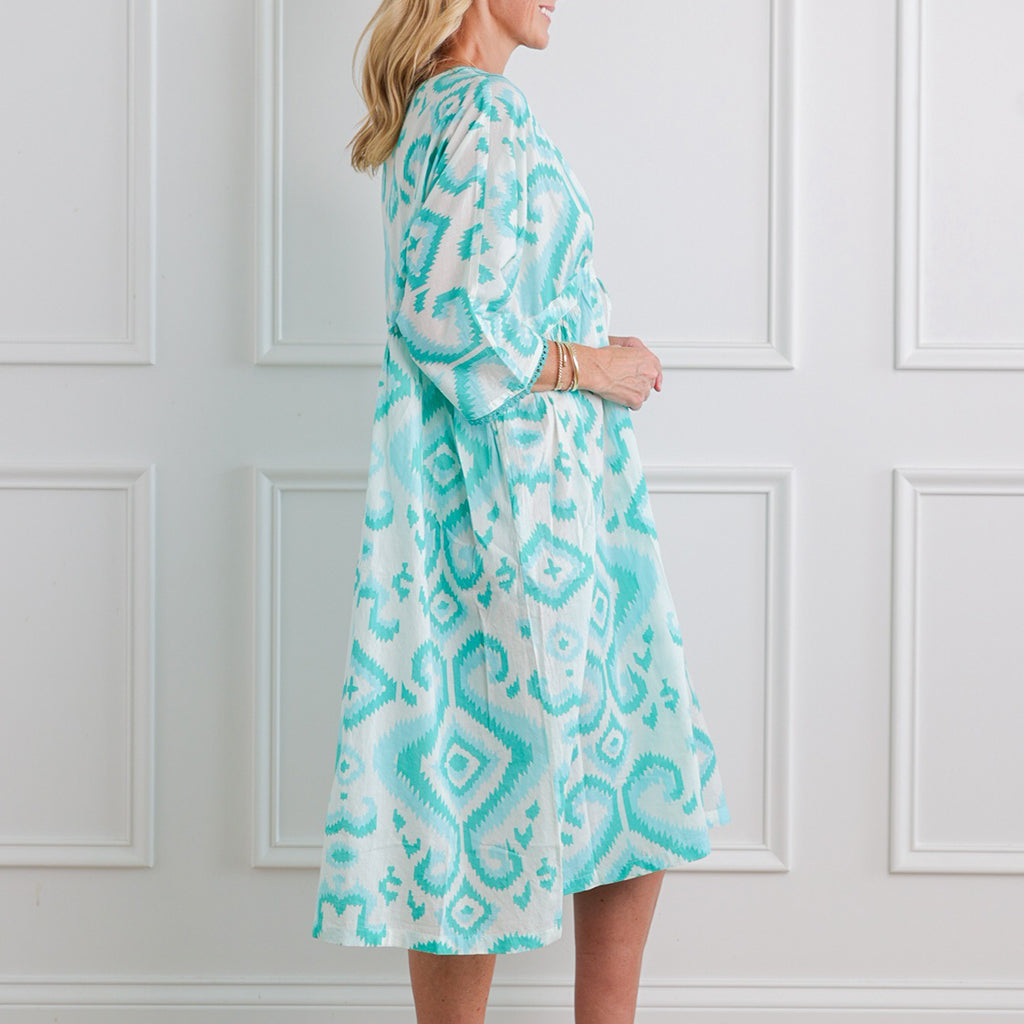 Woman wearing a turquoise and white patterned dress against a white paneled wall.