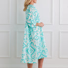 Woman wearing a turquoise and white patterned dress against a white paneled wall.