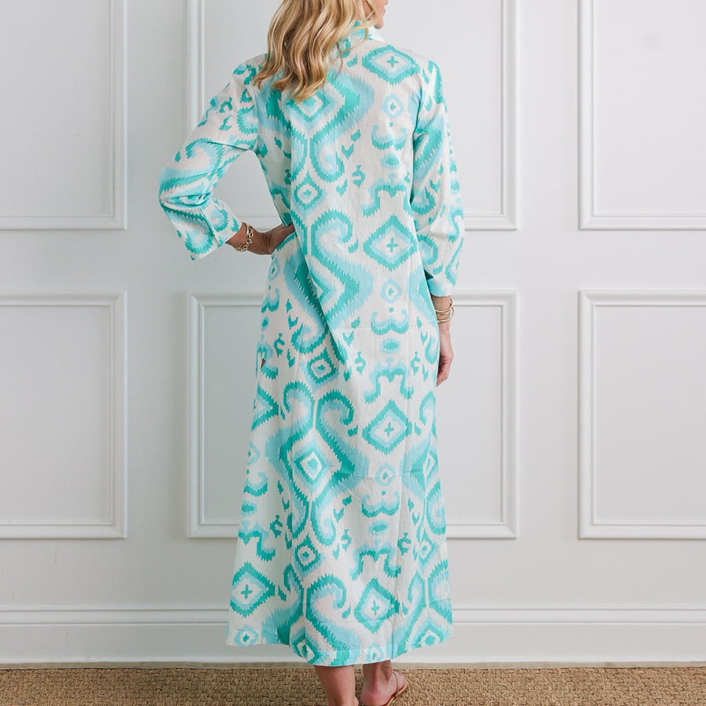 Woman wearing a long, patterned dress with turquoise and white design against a white paneled wall.