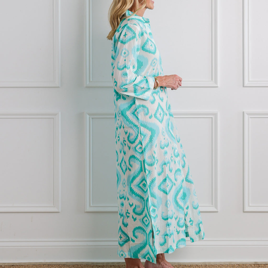 Woman wearing a long, light blue and white patterned dress against a white paneled wall.