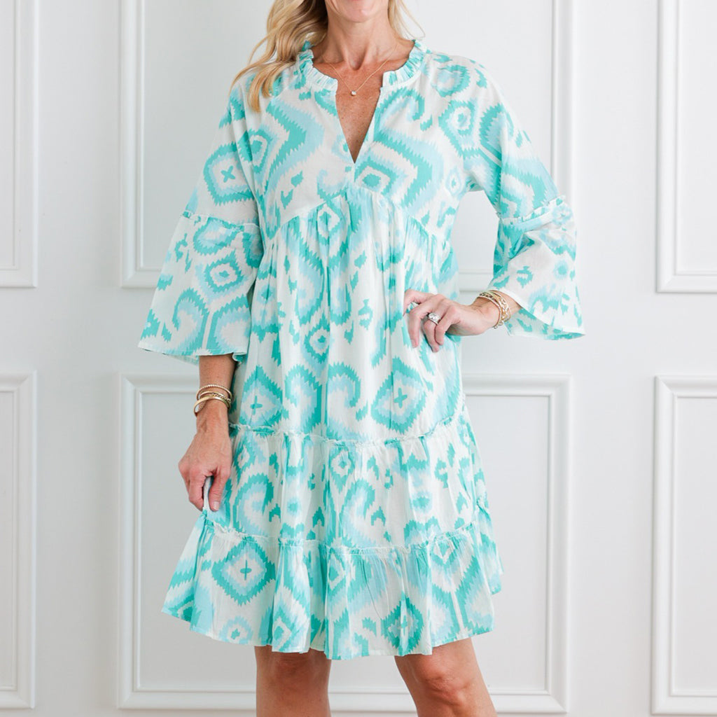 Woman wearing a light blue and white patterned dress against a white paneled wall.