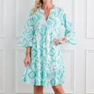 Woman wearing a light blue and white patterned dress against a white paneled wall.