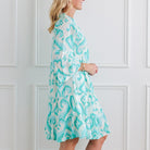 Woman wearing a turquoise and white patterned dress against a white paneled wall.