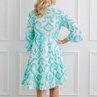 Woman wearing a turquoise patterned dress against a white paneled wall