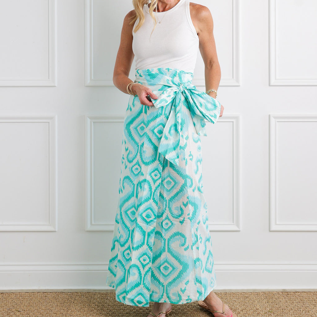 Woman wearing a white sleeveless top and a long turquoise patterned skirt against a white paneled wall.