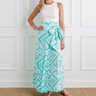 Woman wearing a white sleeveless top and a long turquoise patterned skirt against a white paneled wall.