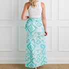 Person wearing a white sleeveless top and turquoise patterned skirt against a white paneled wall.