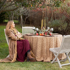 Woman in a fur coat sitting at an outdoor table set for a meal with candles and fruit.