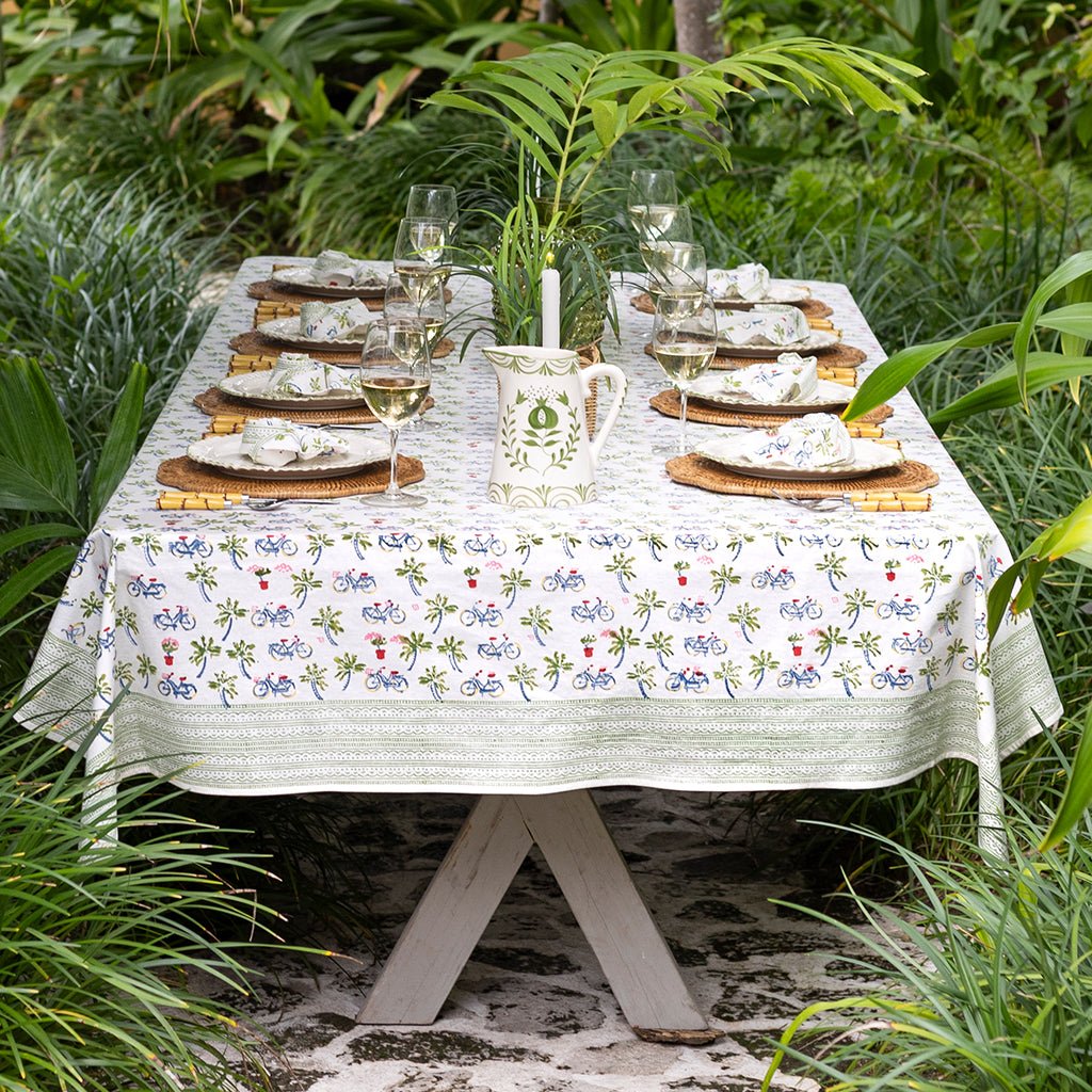 Dining table set with plates, glasses, and a pitcher on a patterned tablecloth outdoors.