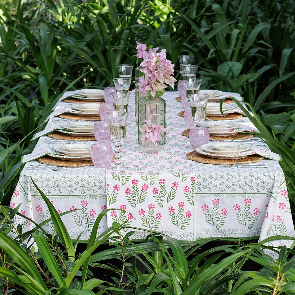 Dining table set with floral tablecloth, plates, glasses, and flowers in a garden setting.