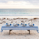 Dining table set with blue tablecloth and white plates on a sandy beach.