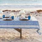 Dining table set with blue and white tablecloth, glasses, and candles on a sandy beach.