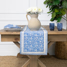 Wooden table with a blue patterned table runner, white flowers in a vase, and blue cups.
