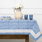Blue patterned tablecloth on a wooden table with white flowers and blue glasses.