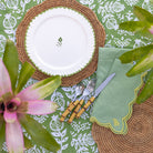Table setting with a white plate, woven placemat, and green napkin on a patterned tablecloth.