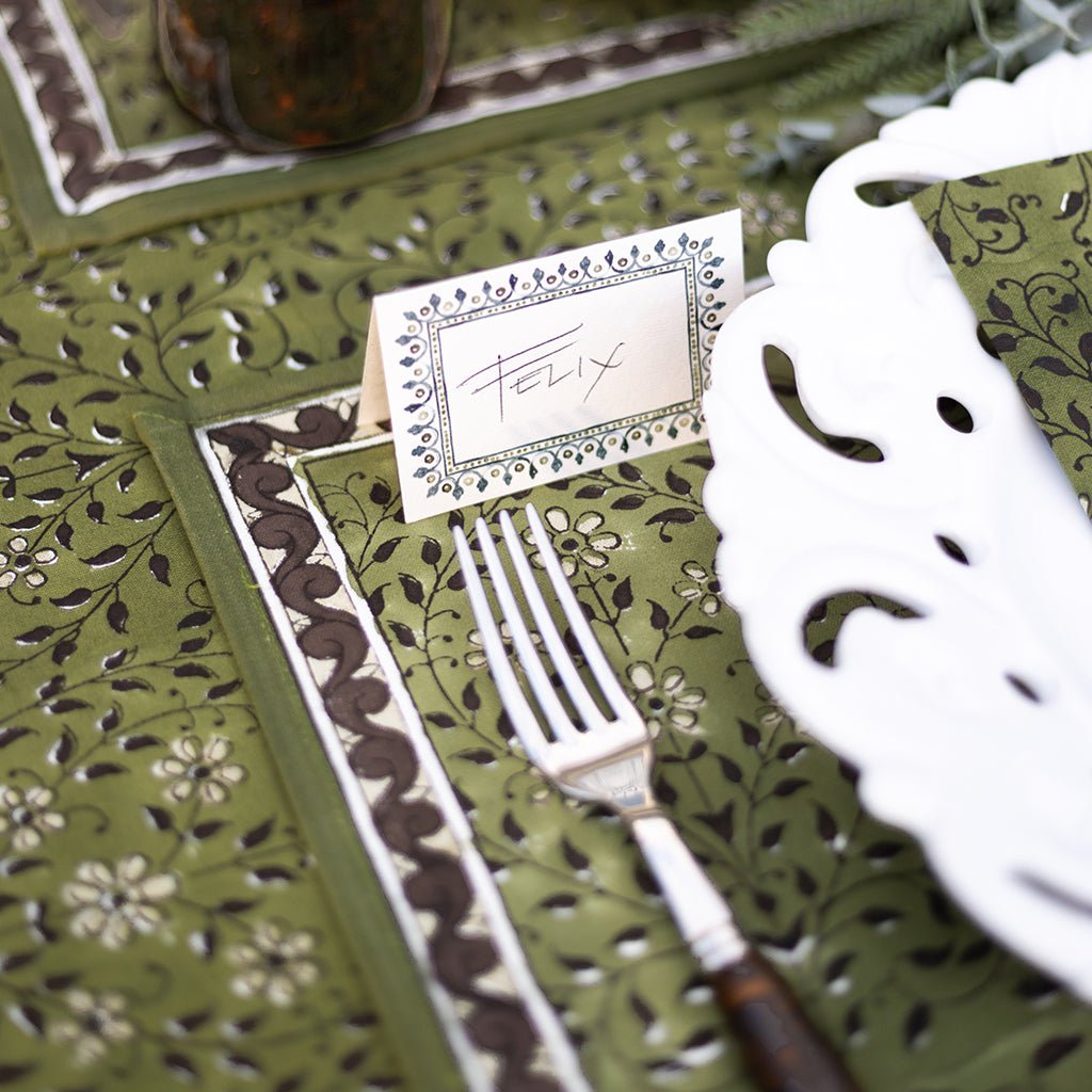 Green patterned tablecloth with a fork, knife, and name card on a decorative setting.