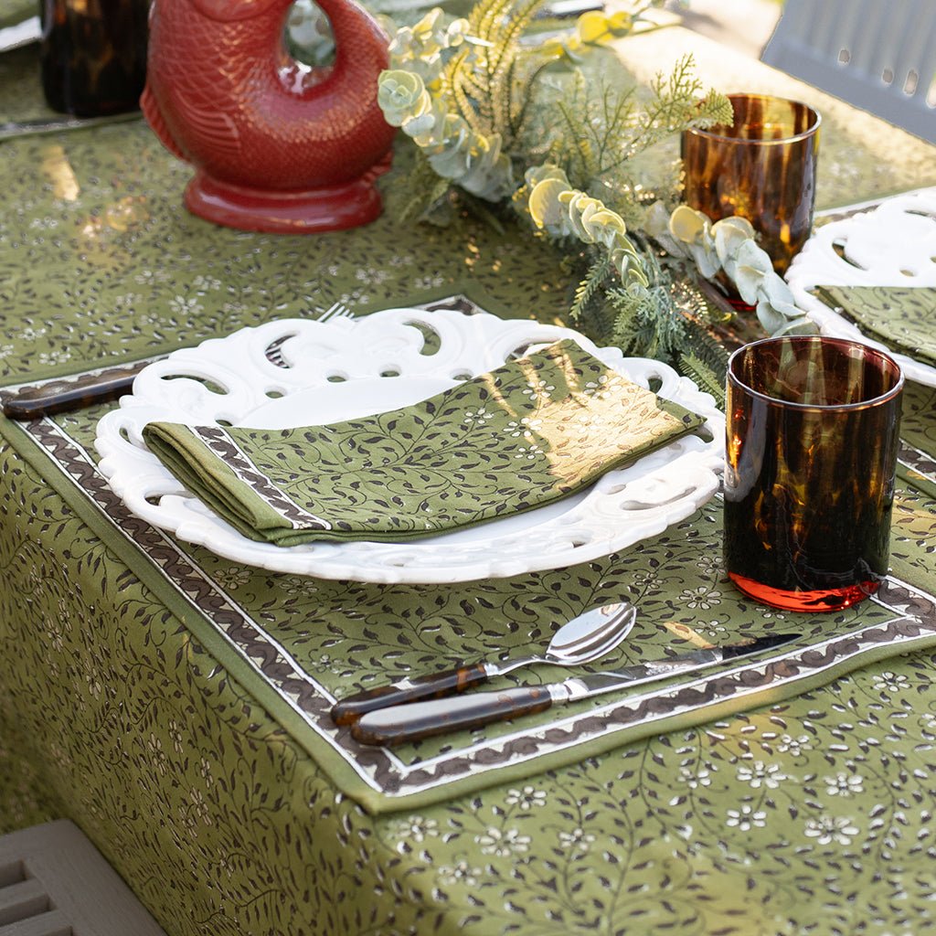 Dining table setting with green placemat, napkin, and tortoiseshell cutlery on a patterned tablecloth.
