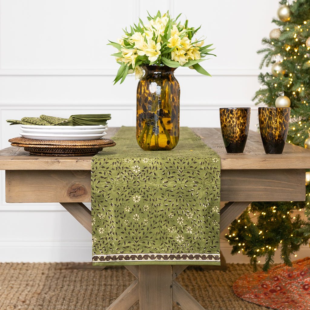 Decorative table setting with a green table runner, vase with flowers, and candles on a wooden table.