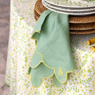Green embroidered napkin hanging on rattan placemats with plates underneath on a patterned tablecloth.