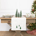 Wooden table with Christmas decor including a white towel with tree designs, mugs, and a decorated Christmas tree.