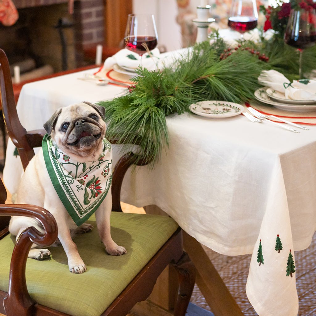 Pug dog wearing a green bandana sitting on a chair at a festively decorated table.