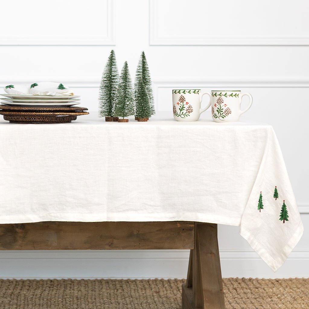 Table setting with a white tablecloth featuring green tree patterns, mugs, and small Christmas trees on a white wall background.